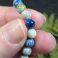 Close-up of a hand holding a bracelet with blue and white beads (azurite, hackmanite, chrysocolla) against a blurred natural background.