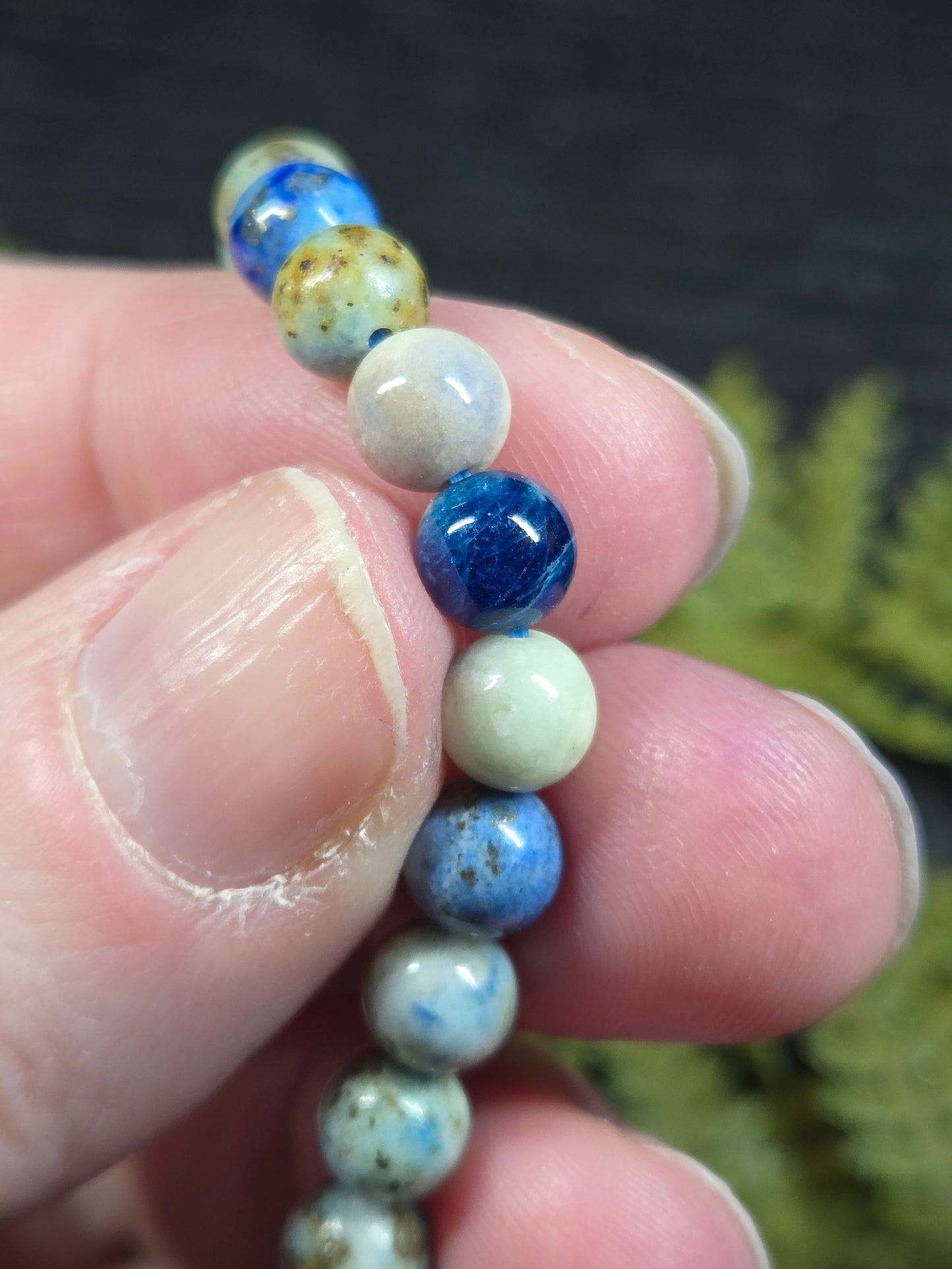 Close-up of a hand holding a bracelet with blue and white beads (azurite, hackmanite, chrysocolla) against a blurred natural background.