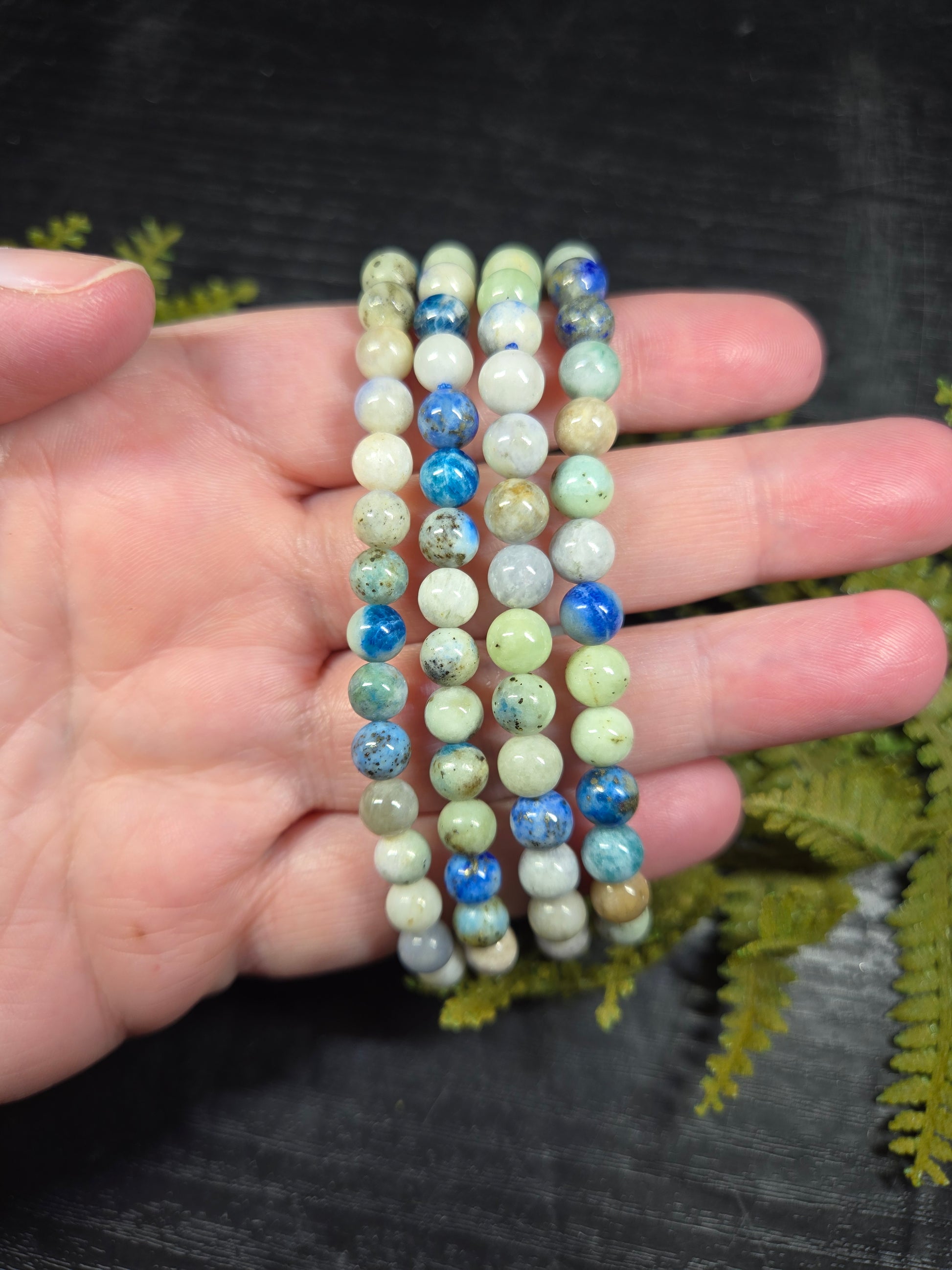 Close-up of a hand holding bracelets with blue and white beads (azurite, hackmanite, chrysocolla) against a dark background.