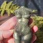 pyrite feminine goddess body carving in sunlight surrounded by green ferns against a black slate background. 