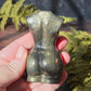 A hand holding a pyrite feminine goddess body carving in sunlight surrounded by green ferns against a black slate background. 