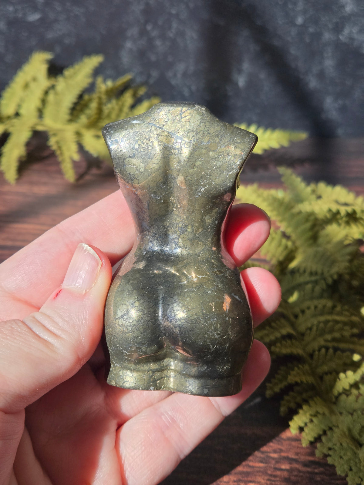 A hand holding a pyrite feminine goddess body carving in sunlight surrounded by green ferns against a black slate background. 