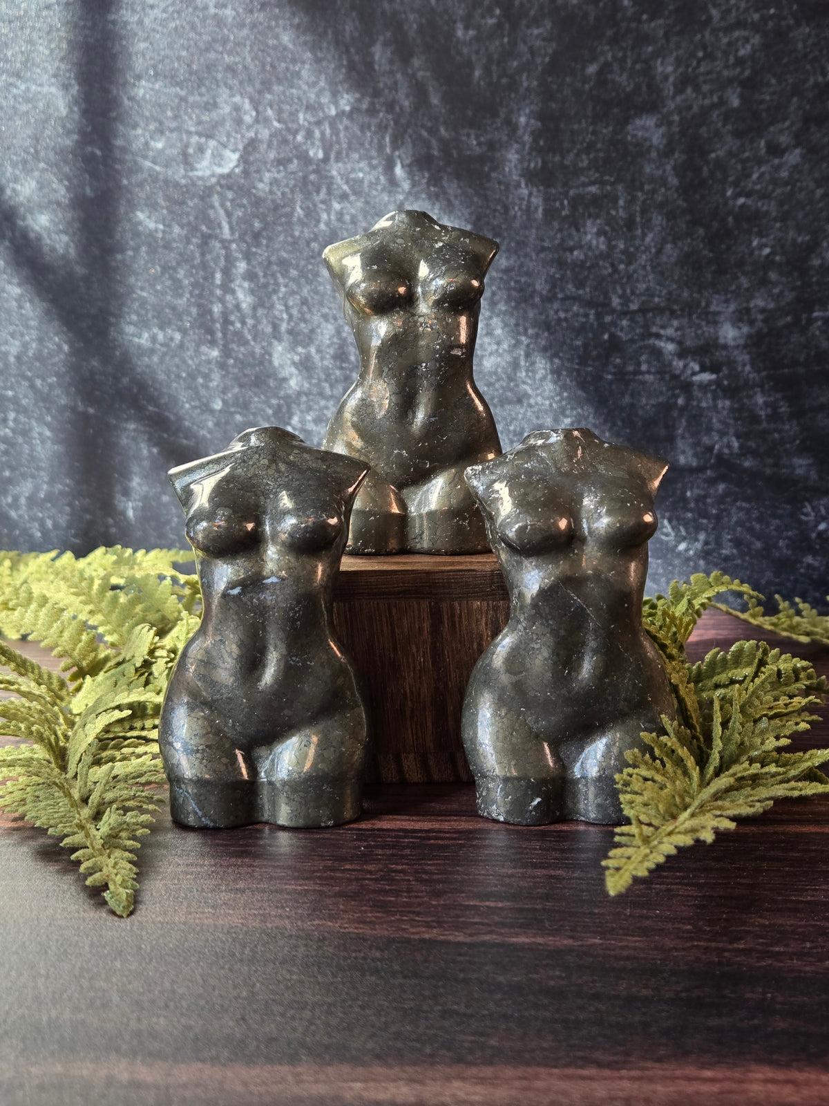 Three pyrite feminine goddess body carvings surrounded by green ferns against a black slate background. 