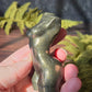 pyrite feminine goddess body carving in sunlight surrounded by green ferns against a black slate background. 