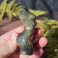 A hand holding a pyrite feminine goddess body carving in sunlight surrounded by green ferns against a black slate background. 